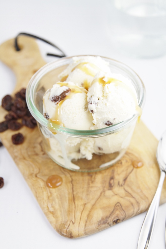 Rum Raisin Ice Cream with Caramel Sauce in small glass jar sitting on cutting board. Raisins and spoon to sides. 