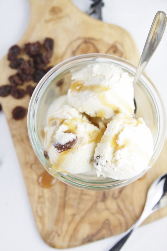 Rum Raisin Ice Cream with Caramel Sauce in glass jar with spoon sitting on cutting board. Pile of raisins behind.