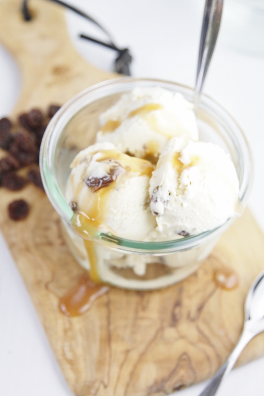 Rum Raisin Ice Cream with Caramel Sauce in glass jar with spoon sitting on cutting board. Pile of raisins to side. 