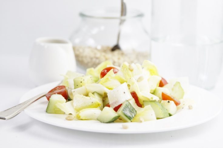 endive tomato salad in white bowl with fork. Glass jar of pine nuts in background.