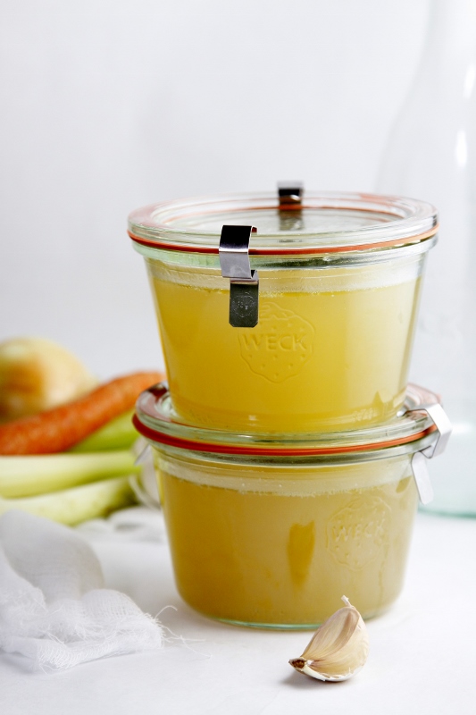 2 glass jars of chicken stock stacked on top of each other. Carrots, celery visible in background.