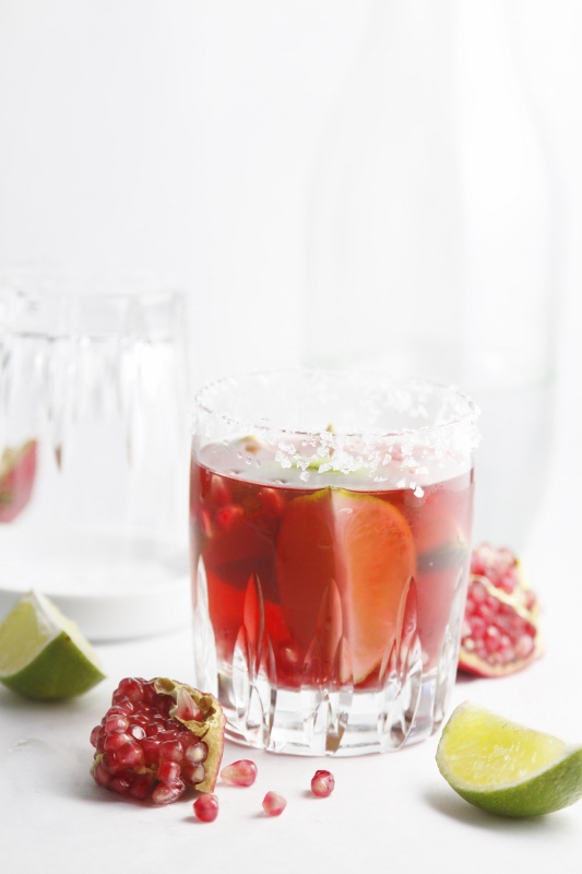 Pomegranate Lime Margarita in glass with lime wedges and pomegrantes scattered around.