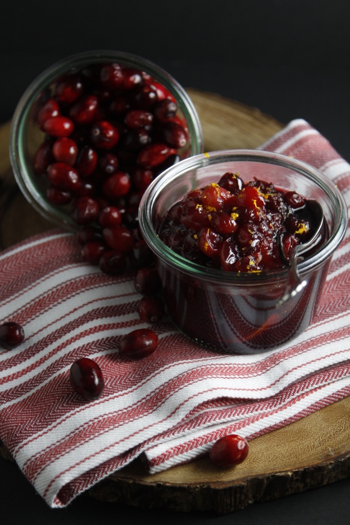 Cranberry Citrus Chutney in glass jar on striped kitchen towel. Glass jar of cranberries tipping over behind chutney.