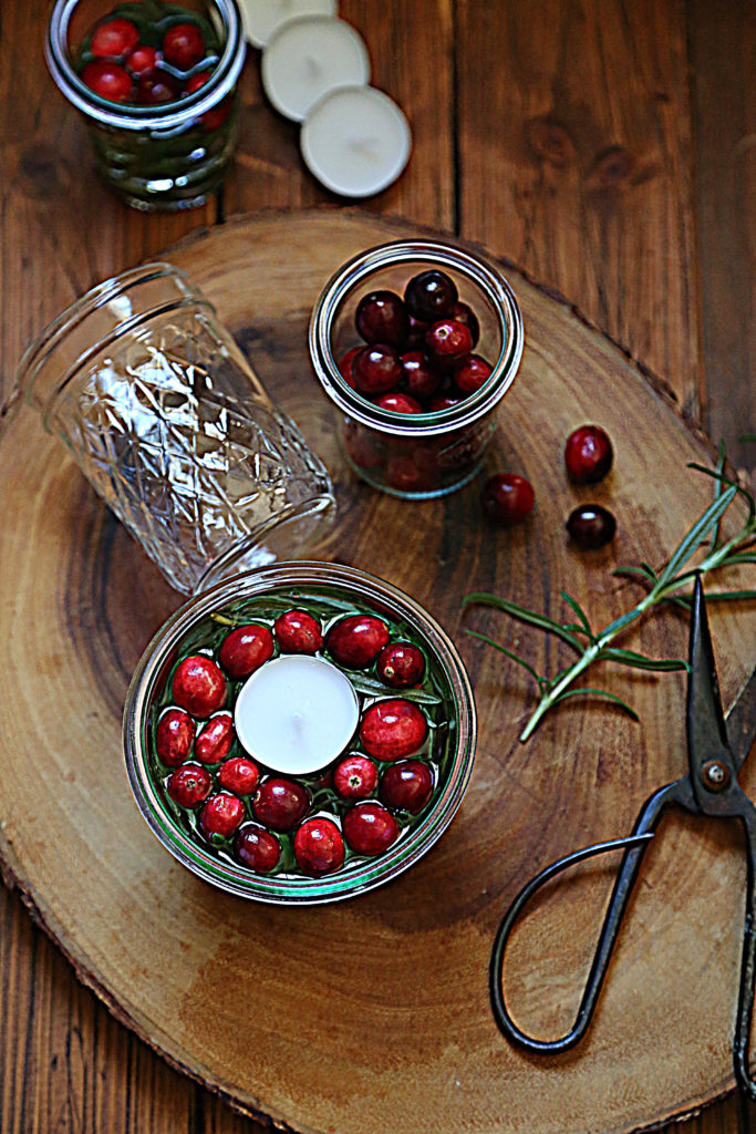 glass jar with floating candle, glass jar of cranberries, scissors and sprig of rosemary, empty glass on side surrounding candle. 