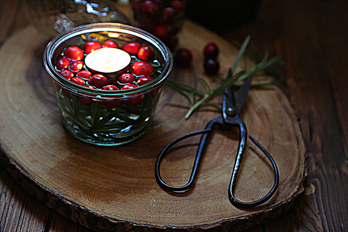 floating candle in glass jar on wood trivet. Scissors, rosemary and cranberries to side. 