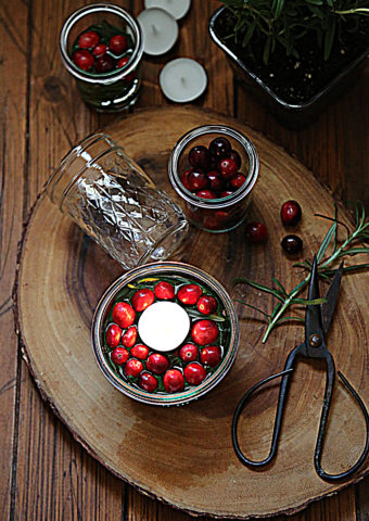 floating candle with cranberries, glass jar of cranberries, glass jar on side, scissors with sprig of rosemary all on wood trivet.