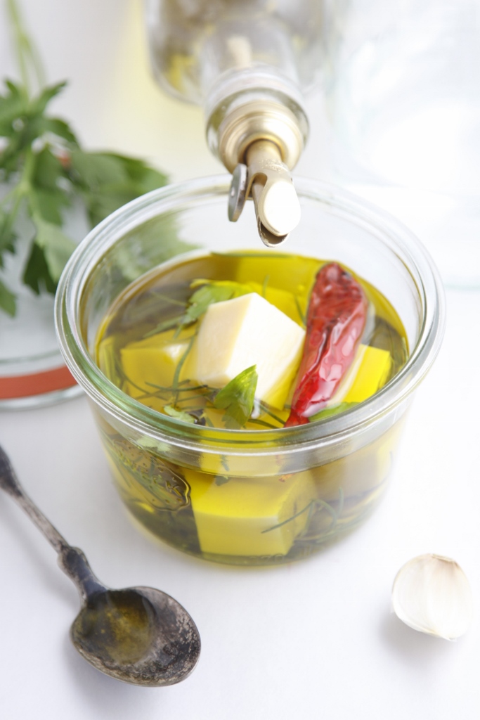 small glass jar of herb marinated cheese cubes. Jar of olive oil resting on side of jar. Spoon, fresh herbs to side. 