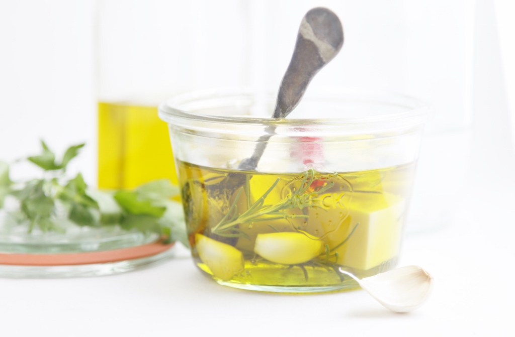Herb Marinated Edam Cheese in glass jar with spoon. Lid with fresh herbs and olive oil jar to side.