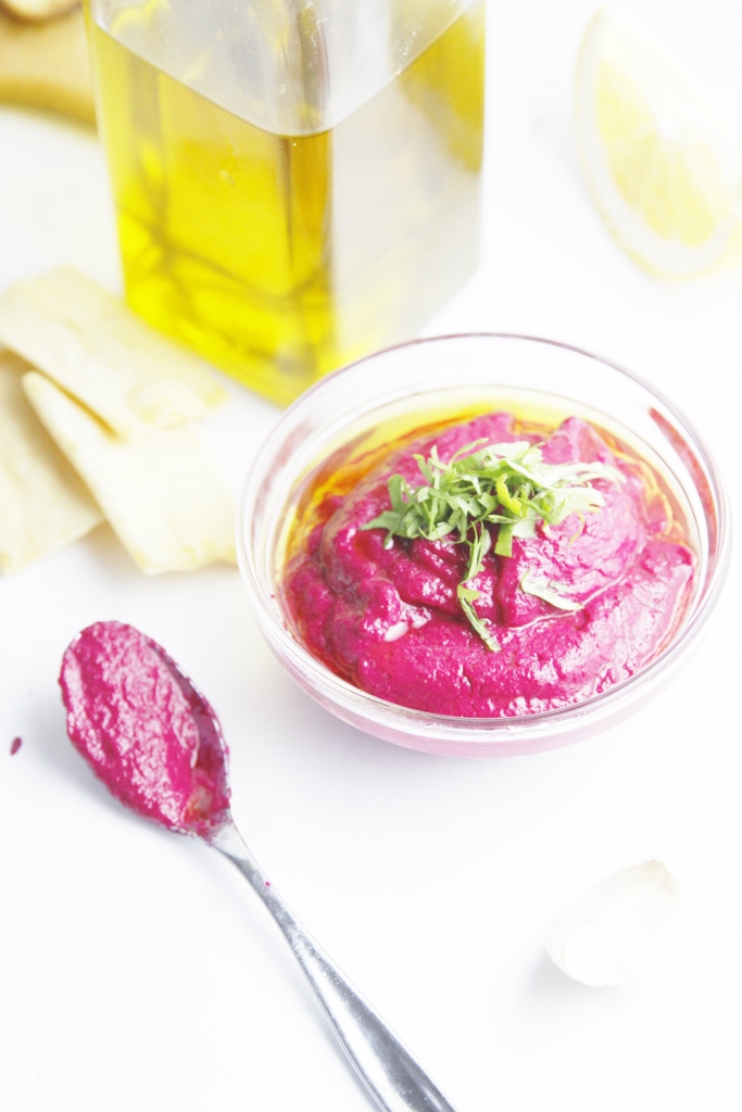 Roasted Beet Hummus in small glass bowl. Spoon with hummus to left. Glass jar of olive oil in background.