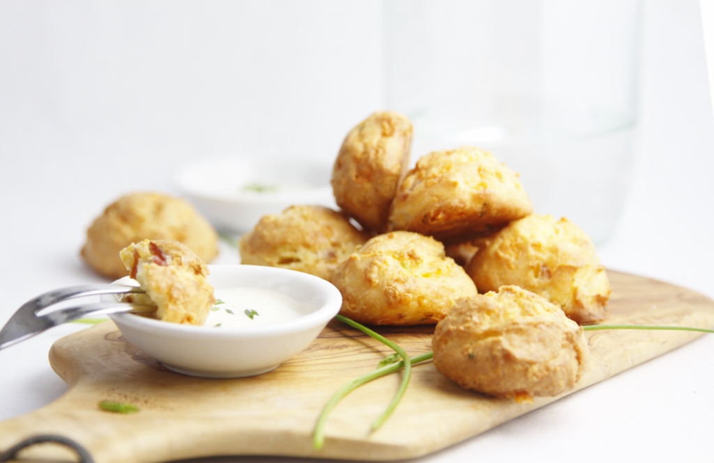 cutting board with stack of Gougeres and small white bowl with dipping sauce. Chives scattered around.