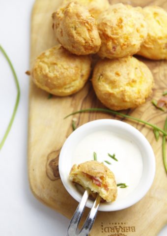stack of Gougeres on cutting board with small white bowl of dipping sauce. Chives scattered around.