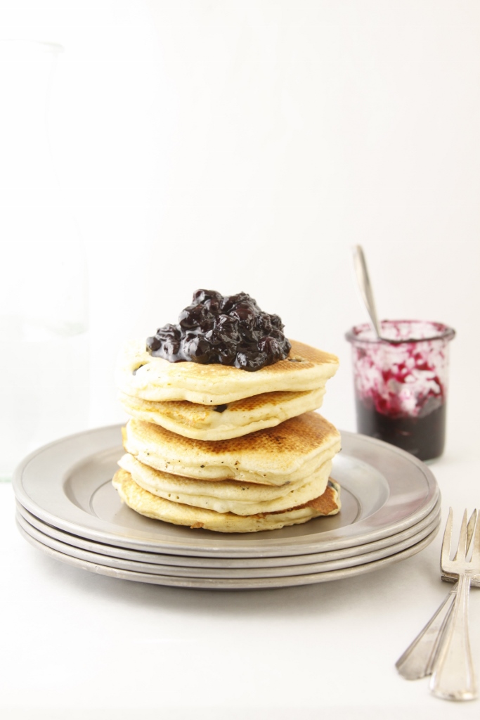 stack of pancakes topped with blueberry compote on metal plates. Glass jar of compote with spoon behind.