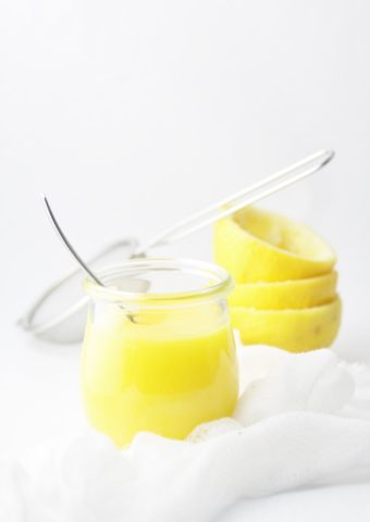 lemon curd in small glass jar with spoon. Lemons behind.