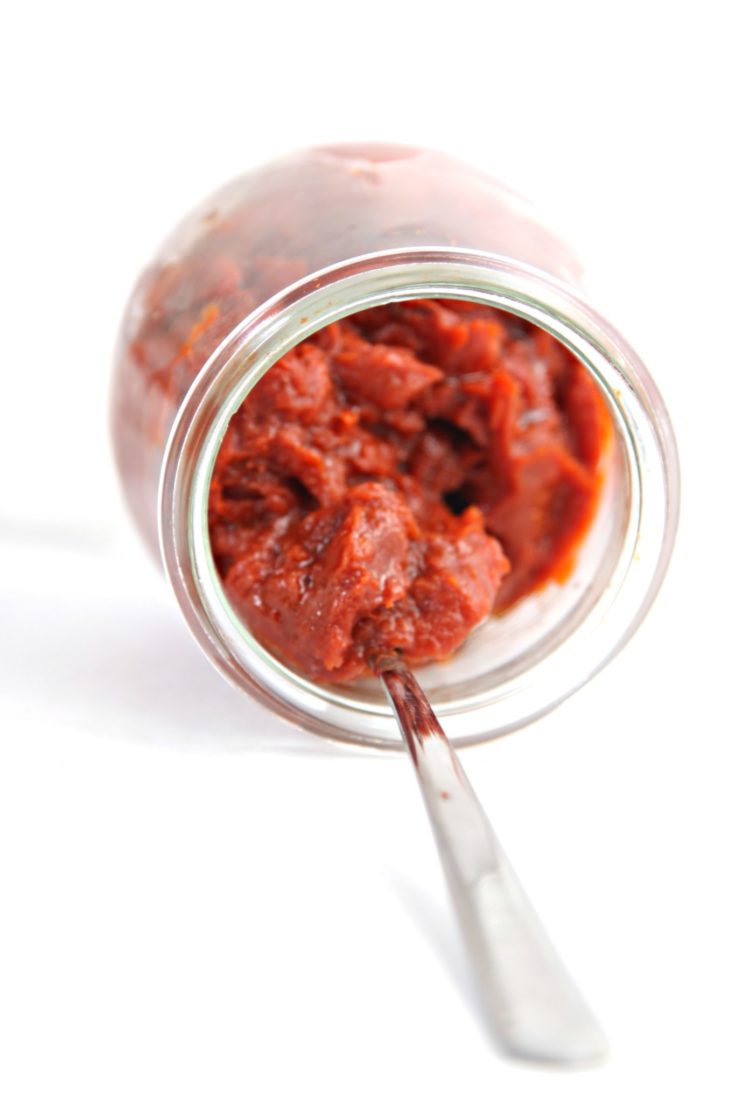 small glass jar on side full of homemade tomato paste. Small silver spoon inside jar.