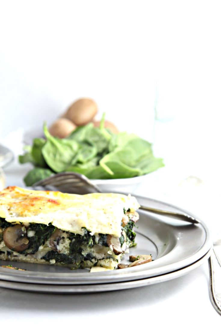 slice of spinach mushroom lasagna on silver plate with fork. Bowl of mushrooms and spinach behind.
