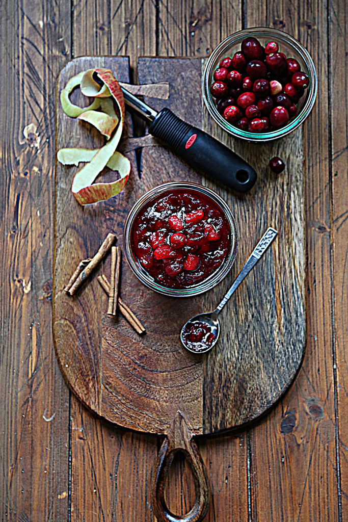 cutting board with glass jar of cranberry sauce, glass jar of cranberries, cinnamon sticks, spoon with sauce, apple peel and peeler. 