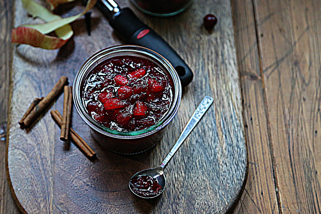 glass jar of cranberry sauce, spoon with sauce, cinnamon sticks, apple peel and peeler on cutting board. 
