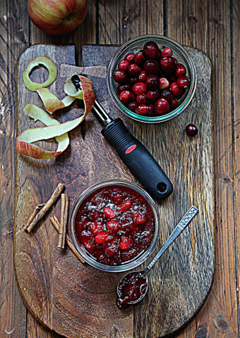 cutting board with apple peel, peeler, cinnamon sticks, jar of cranberries and jar of cranberry sauce.