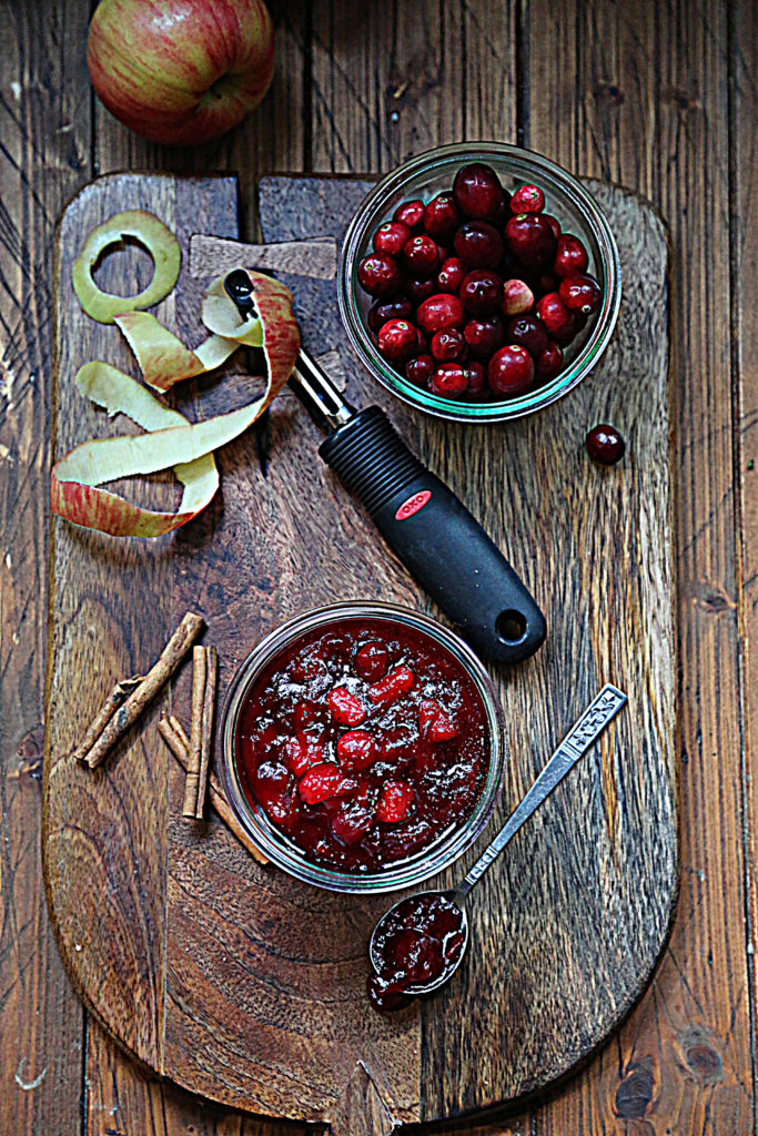 cutting board with apple peel, peeler, cinnamon sticks, jar of cranberries and jar of cranberry sauce.