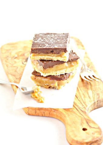 cutting board with 3 caramel chocolate shortbread bars stacked on each other. Fork to side.