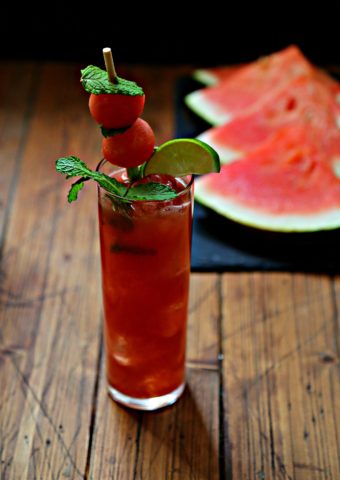 tall glass of watermelon mojito garnished with melon balls, lime wedge and mint. Slices of watermelon in background.