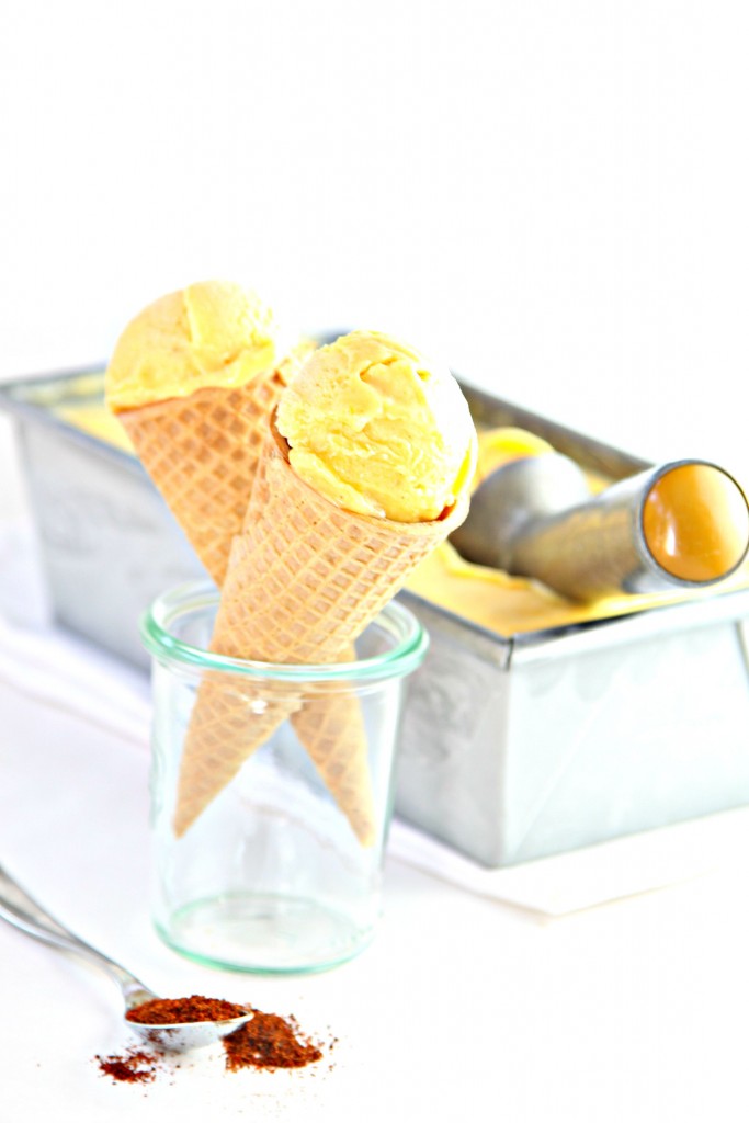 loaf pan of Mango Lime Chili Ice Cream with ice cream scooper. Small glass jar with 2 ice cream cones standing up.