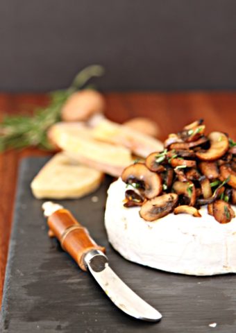 Savory Baked Brie with Crispy Mushrooms on a slate board. Cheese knife to side. Mushrooms and rosemary behind.