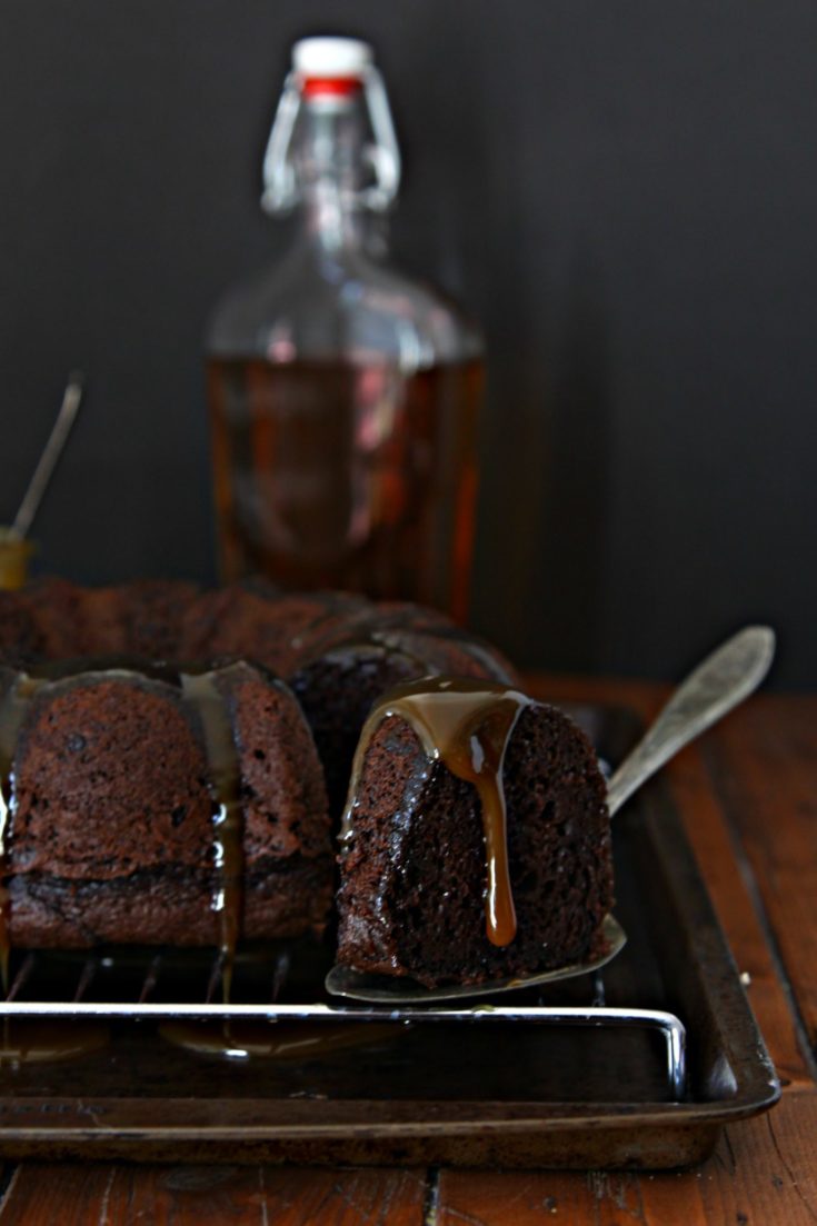 chocolate bundt cake on cooling rack. Slice of cake being pulled out. Bottle of bourbon behind.