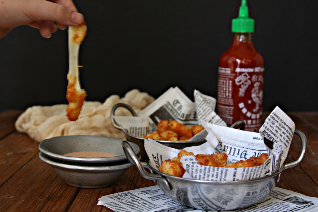 Two small metal bowls lined with newspaper filled with Fried Cheese Curds. Bottle of sriracha in background. Hand holding cheese curd above small metal bowl with dipping sauce. 