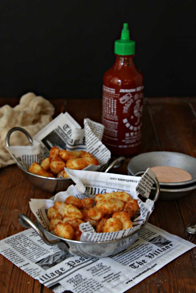 Two small silver bowls with handles lined with newspaper filled with Fried Cheese Curds. Bottle of sriracha in background. Small silver bowl of sauce to right. 