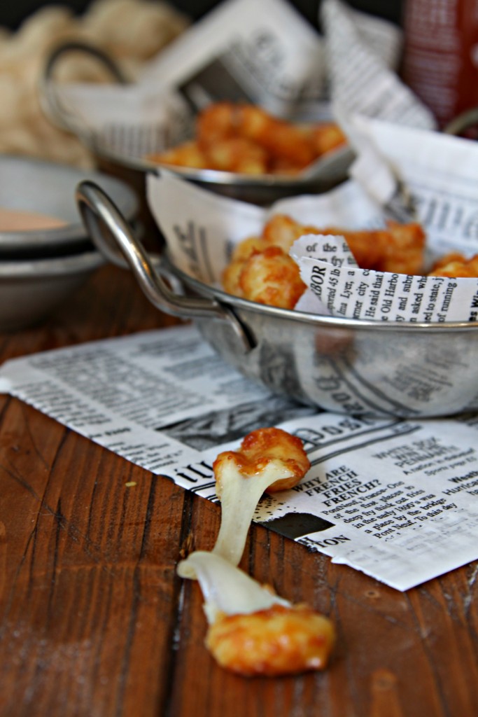 Two small silver bowls with handles lined with newspaper filled with Fried Cheese Curds. One cheese curd with cheese pull in front of bowls. 