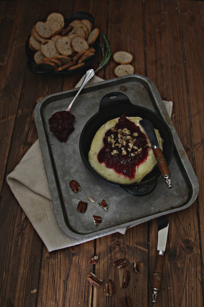 Baked Brie With Cranberries in cast iron skillet sitting on baking sheet. Serving spoon , knife and pecans scattered around. 