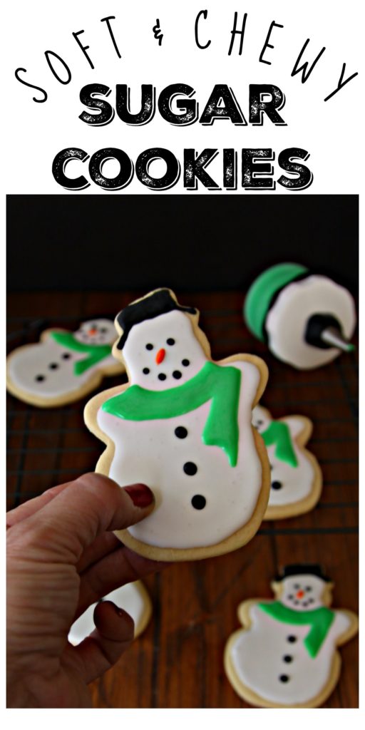 Pinterest image. Hand holding Snowman Sugar Cookie. Additional cookies in background with applicator. Text overlay that reads Soft & Chewy Sugar Cookies. 