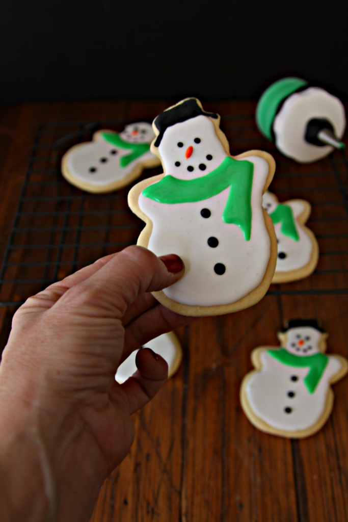 hand holding a Snowman sugar cookie. Snowman sugar cookies in background on cooling tray. Icing applicator on tray. 
