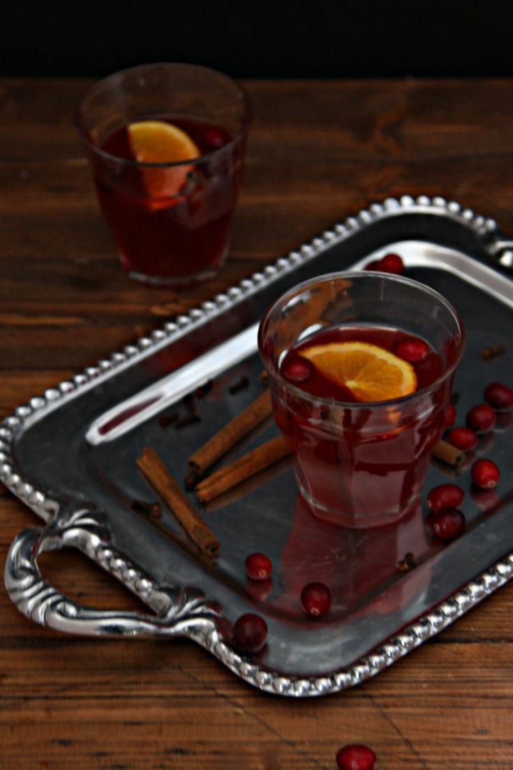 silver tray with glass of punch. fresh cranberries and cinnamon sticks scattered around glass. Second glass of punch in background.