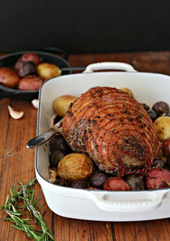 Herb Roasted Leg of Lamb in white baking dish surrounded by potatoes.