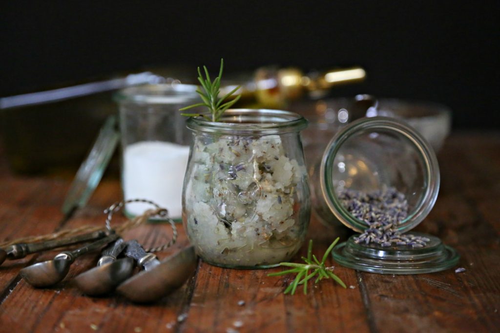 Small glass jar of Rosemary Lavender Sugar Scrub in center. Small glass jar of lavender to right spilling out. Small glass jar of sugar in background with blurred bottle of olive oil. Measuring spoons in front.