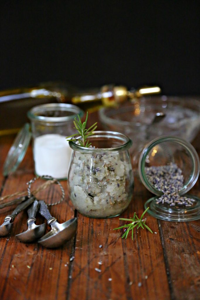 Small glass jar of homemade Rosemary Lavender Sugar Scrub. Measuring spoons, small glass jar of sugar, small glass jar of lavender tipped over in background. 