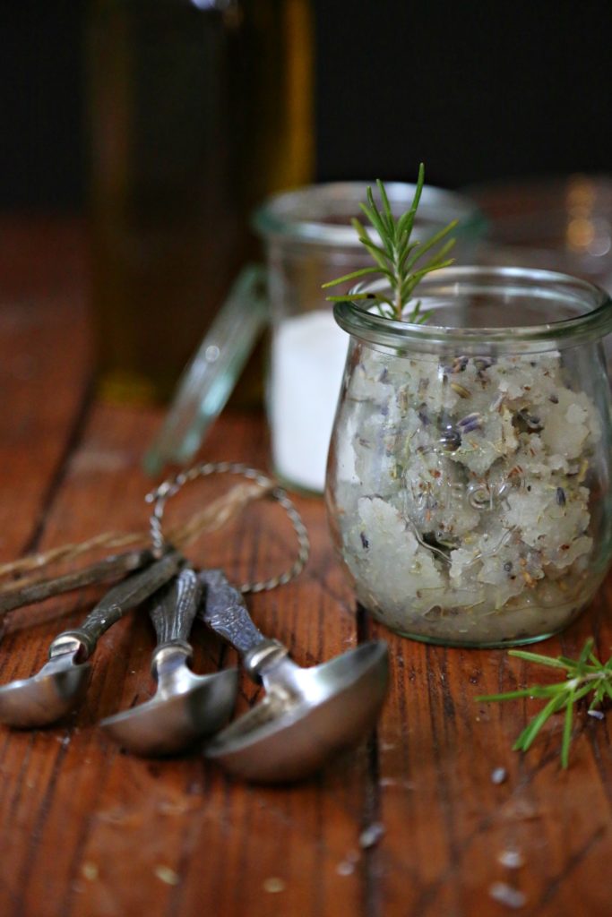 Small glass jar of Rosemary Lavender Sugar Scrub. Small glass jar of sugar, bottle of olive oil in background. Measuring spoons in front. 