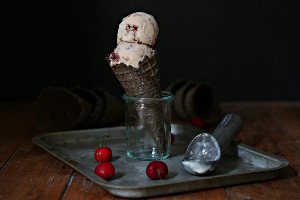 Two scoops of Roasted Cherry Bourbon Ice Cream in chocolate cone standing in glass jar on baking sheet. Cherries and ice cream scoop around glass jar. 