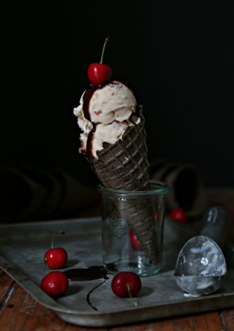 Roasted Cherry Bourbon Ice Cream in chocolate ice cream cone standing in a small glass jar on a baking sheet. Cherries scattered around jar.