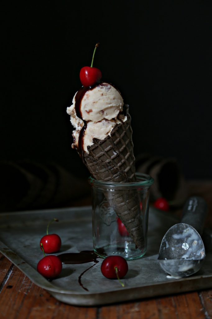 Two scoops of roasted Cherry Bourbon Ice Cream in chocolate cone, standing in small glass jar on baking sheet. Cherries scattered around. Ice cream scoop to right. 
