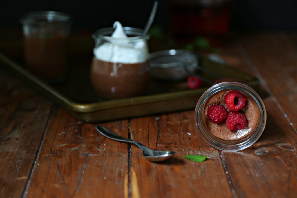 2 small glass jars of Chocolate Bourbon Mousse on baking tray with metal sifter. One small glass jar of mouse on it's side in front garnished with raspberries. Small spoon to side of jar. 