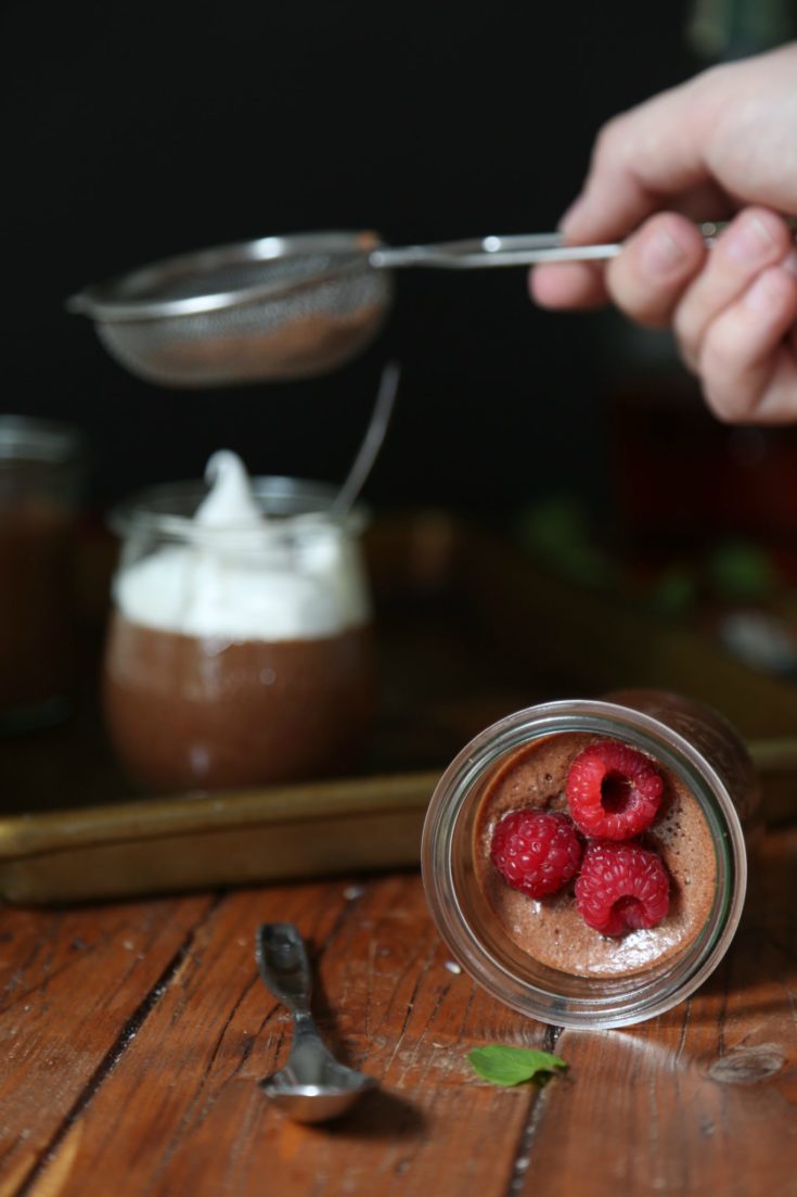 Chocolate Bourbon Mousse in small glass jars. Garnished with raspberries. Sitting on baking sheet. Sieve of chocolate sprinkling on top.