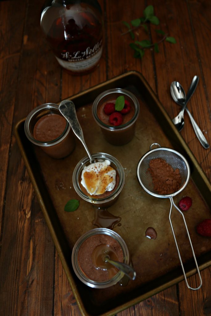 4 small jars of Chocolate Bourbon Mousse on a baking tray. Small metal sifter with cocoa powder to side. Bottle of bourbon in rear of photo. Small spoons to right of baking tray. 