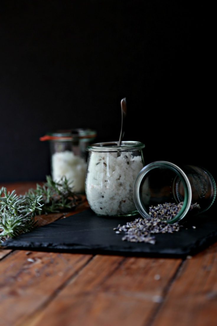 DIY Lavender Orange Sugar Scrub in small glass jars. Fresh rosemary sprigs to side, plus jar of coconut oil with spoon.