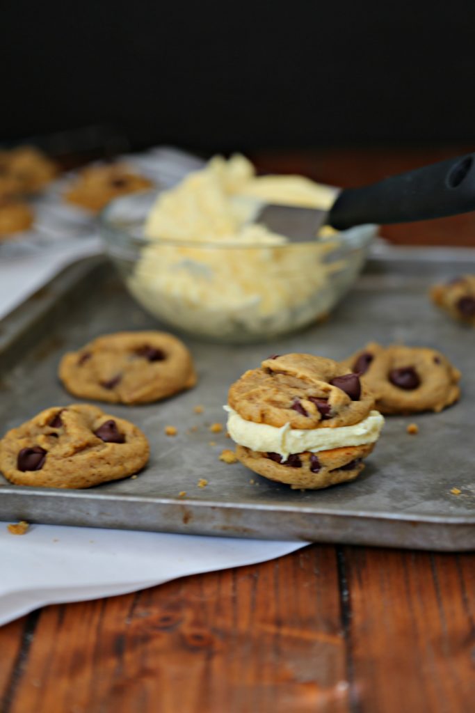 Pumpkin Chocolate Chip Cookies on baking sheet with a small glass bowl of frosting with spatula. 