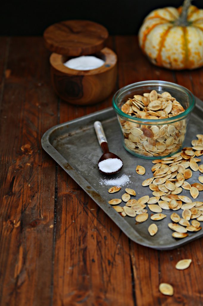 glass jar of roasted pumpkin seeds on baking tray with scattered seeds and spoon of salt. Salt cellar in background with pumpkin.