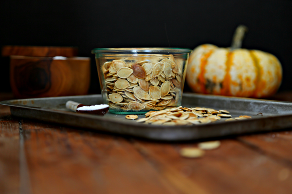 roasted pumpkin seeds in glass jar on baking sheet. Pumpkin and salt cellar in background.