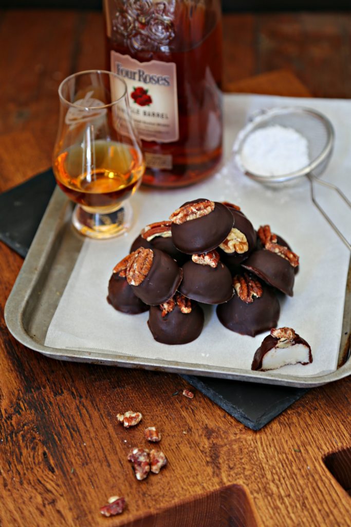 Bourbon Balls stacked on baking tray with bottle and glass of bourbon, small metal sifter with powdered sugar. 